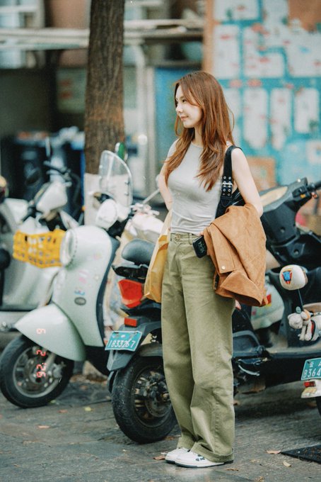 The image features a woman standing next to two motorcycles, one white and one yellow. She has her handbag over her shoulder and appears to be wearing a gray shirt. The background of the photo shows a building with blue walls, which adds an urban feel to the scene. There is also text in the image that reads "I closed to survive that is how you stay in this game. So did I. ... That is how you stay in this game. ... That is how you stay in this game." This message seems to be related to the woman's motorcycle experience, as she has chosen to ride a motorcycle instead of using public transportation or walking.
