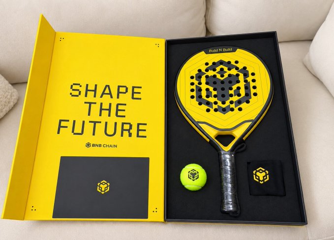 An open yellow and black presentation box containing a yellow padel racket branded 'BNB CHAIN' with the slogan 'SHAPE THE FUTURE', along with a yellow ball and accessories.