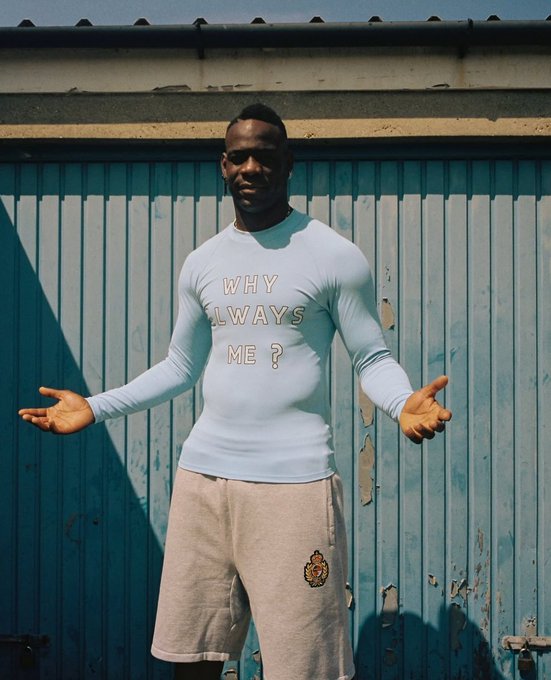 A man stands with his arms outstretched in front of a blue corrugated metal door, wearing a light blue long-sleeved shirt that reads 'WHY ALWAYS ME?' and gray shorts.