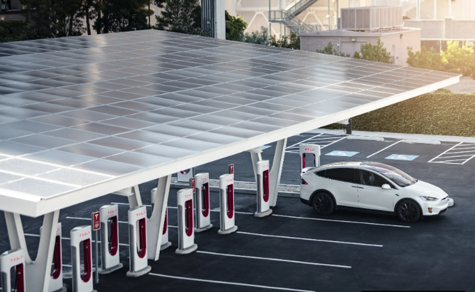 A Tesla electric vehicle charging station with multiple stalls under a large solar panel canopy, with a white car parked at one of the charging spots.