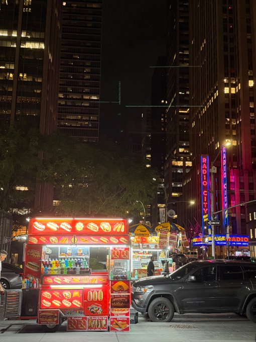 A vibrant nighttime street scene in New York City, featuring illuminated "HOT DOG," "SAUSAGE," and "CHICKEN" food carts in the foreground, with the iconic "RADIO CITY" neon sign and "Radio City Music Hall" marquee glowing in the background beside a parked SUV.