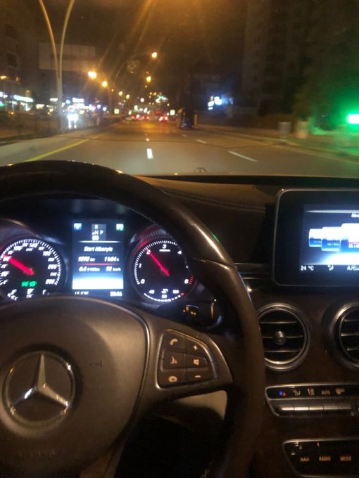 A nighttime view from the driver's seat of a Mercedes-Benz shows the illuminated dashboard playing 'Don't Worry' and a city street ahead.