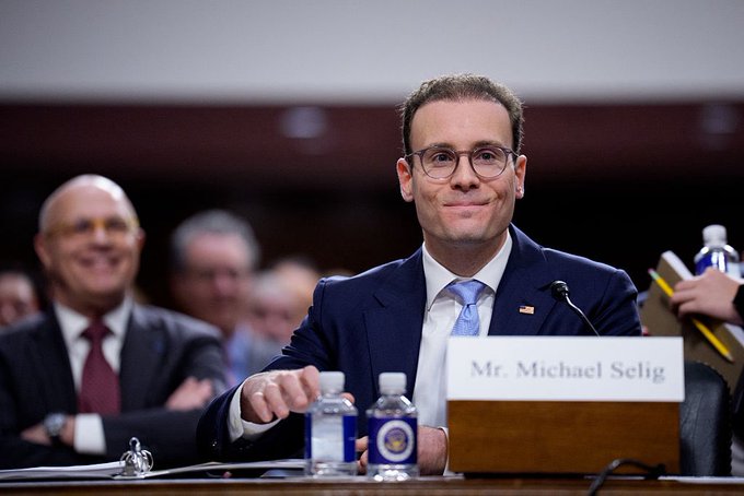 A man wearing glasses, a blue suit, and a light blue tie smiles at a hearing, with a nameplate reading 'Mr. Michael Selig' in front of him.