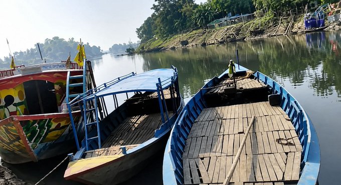 A photo taken from the deck of a boat looking forward, showing two blue wooden passenger boats docked side-by-side on a calm river, with forested banks and traditional buildings visible in the background under a hazy sky.