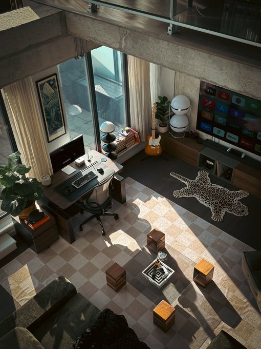 A loft-style room with floor-to-ceiling windows features a gaming/computer desk setup, a guitar, and a large TV displaying an Apple TV interface with visible app icons including Apple TV+, Netflix, Disney+, YouTube, and Prime Video. The space is decorated with a leopard print rug, potted plants, and wooden cube seating arranged around a central coffee table on a checkered rug.