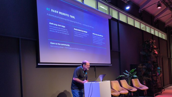 A man standing at a podium giving a presentation in front of a projection screen titled '02 DASH WEBSITE TOOL,' which describes a decentralized publishing tool and its features.