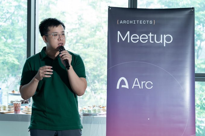 A man in a green polo shirt speaking into a microphone at an {ARCHITECTS} Meetup event, standing next to a banner displaying the 'Arc' logo.