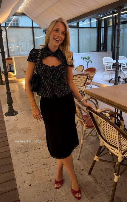 A smiling blonde woman in a black polka-dot top and skirt stands in an outdoor café setting with the caption 'my heart is so full'.