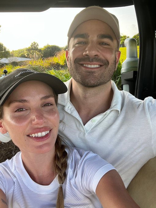 A selfie of a smiling couple wearing caps in what appears to be a vehicle, with the woman wearing a black cap with white text and a braid.