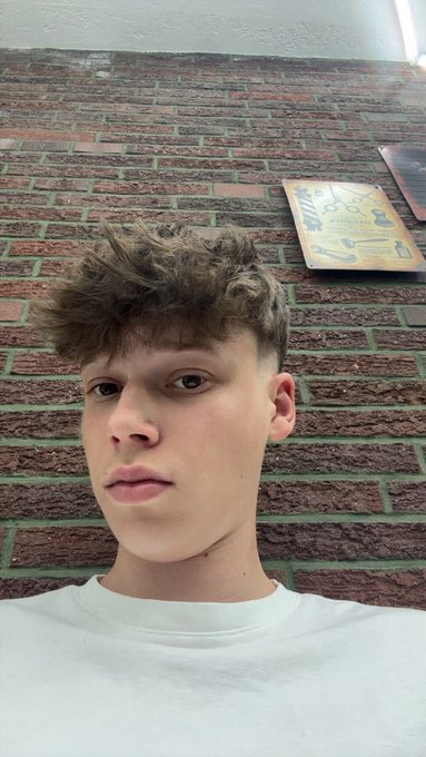 A selfie of a young man with curly brown hair wearing a white t-shirt, standing against a brick wall with a gold-colored plaque visible behind him.