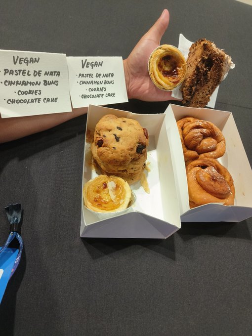 A display of vegan pastries including Pastel de Nata, Cinnamon Buns, Cookies, and Chocolate Cake, with handwritten labels identifying each item.