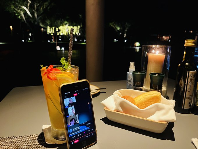 A nighttime dining table scene with a tall cocktail garnished with mint and a straw, a smartphone propped up displaying a video call, a bread basket with a white napkin, a lit candle, and a bottle of Castelli olive oil.