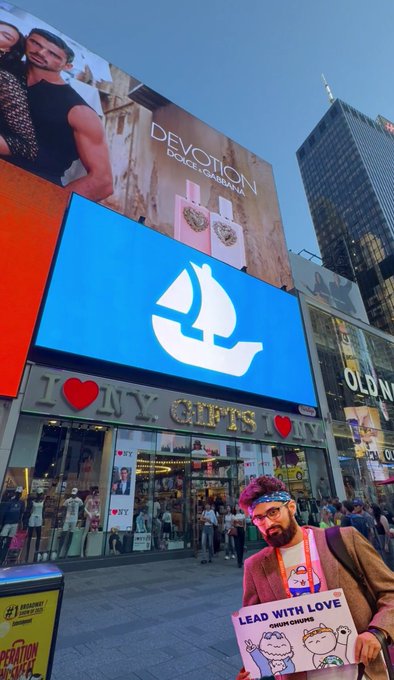 A person holding a sign reading 'LEAD WITH LOVE' from HAIM & CHIENS stands in Times Square near the I♥NY Gifts store and Devotion Dolce & Gabbana billboard.
