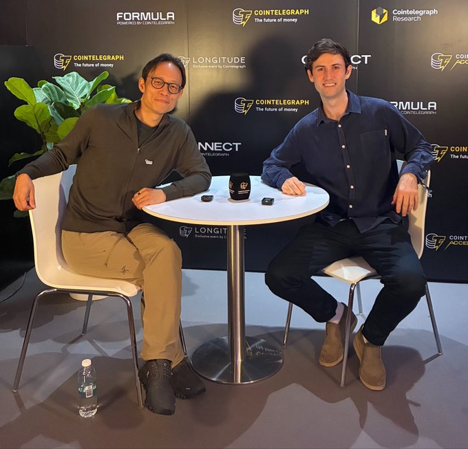 Two men sitting at a table with Cointelegraph and Formula branded mugs and equipment, appearing to record a podcast or interview, with Cointelegraph and Cointelegraph Research logos in the background.