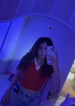 A young woman with long dark hair takes a mirror selfie in a blue-lit bathroom while wearing a red short-sleeved top and denim jeans.