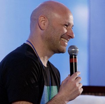 The image shows a bald man wearing a black shirt standing behind a microphone, holding it up and smiling at the camera. The backdrop of the scene appears to be a blue wall, providing a contrasting color to the man's appearance. A caption next to the photo states that it is "eth crash szn?" three years of silence then $121METH moves in one shot, indicating an interesting development happening around this individual at the moment depicted in the image.