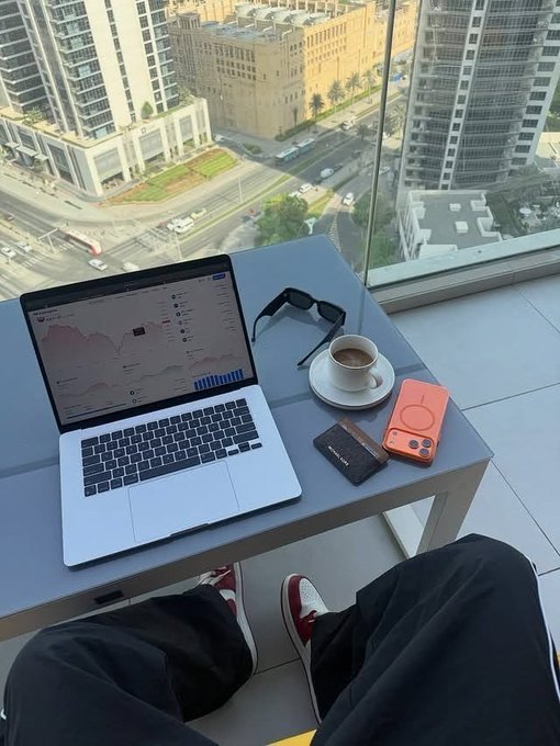 A first-person view of a person working on a MacBook displaying analytics charts on a balcony, with sunglasses, a coffee cup, a black cardholder, and an orange phone accessory on the table overlooking a city.