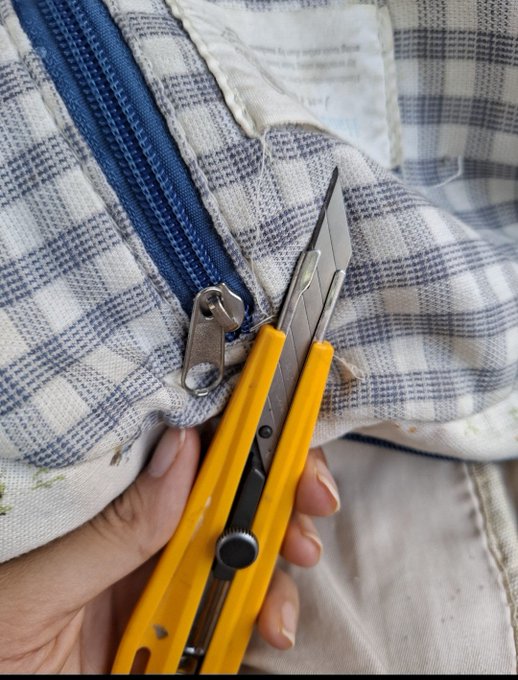 A close-up of a hand holding a yellow utility knife near the blue zipper of a blue and white plaid fabric bag or pouch.