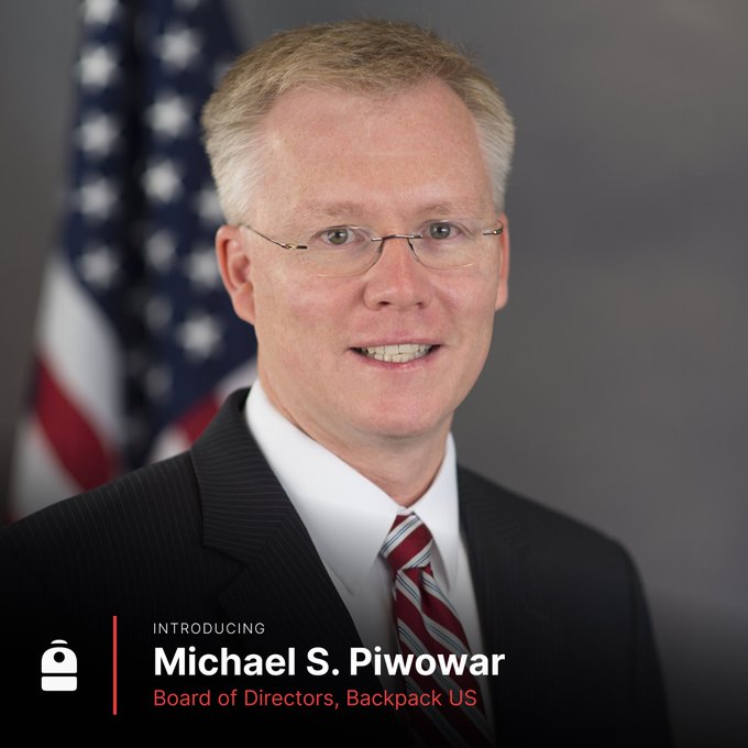 The image features an older man wearing glasses, a suit, and a red tie with white stripes. He has a smile on his face as he poses for the picture. The background of the photo includes an American flag, which suggests that this is likely taken in the United States. In the bottom right corner of the image, there's text that reads "Introducing Michael S. Piwowar Board of Directors Backpack US". This indicates that Michael Piwowar has joined the Backpack US board as a member and is now officially part of their organization.