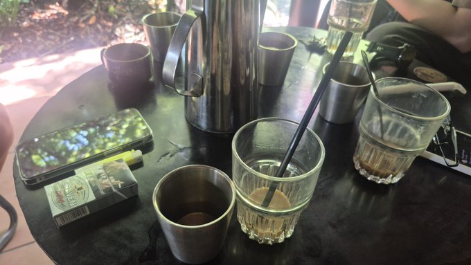 A casual tabletop scene featuring drinking glasses with beverages and black straws, metal cups, a smartphone, and a pack of cloves cigarettes on a dark table.