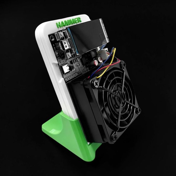 Close-up product photo of a HAMMER-branded electronic device featuring an exposed circuit board, OLED display, USB-C port, a black cooling fan with wire guard, mounted in a white and green 3D-printed stand against a black background.