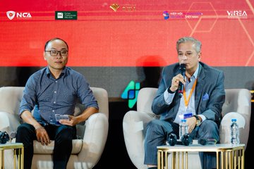 Two men sit on white chairs at a panel during the Vietnam RWA Summit 2026, with banners for NCA, VNX, and VARA visible in the background, as the man on the right speaks into a microphone while the man on the left listens attentively.