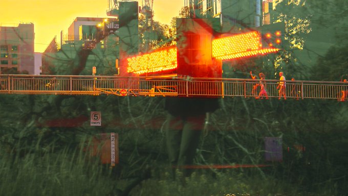 A double-exposure style image showing pedestrians on a bridge overlaid with a digital electronic sign displaying orange LED text.