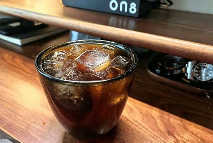 A glass of iced coffee (or iced Americano) sits on a wooden desk beside a watch, with a digital display in the background showing "18:10." The image accompanies the post as a casual morning drink snapshot — though the visible time (6:10 PM) is a playful contrast to the "#每日咖啡简报" (Daily Coffee Briefing) theme of the Bitcoin news.