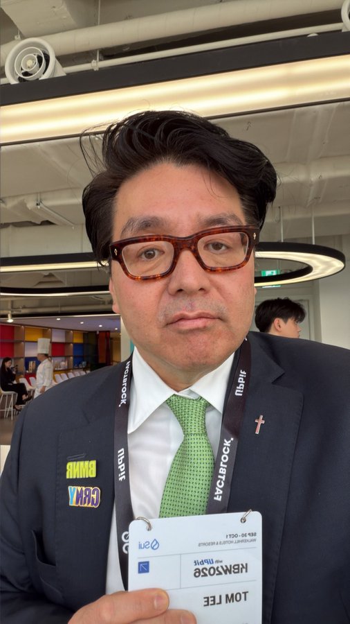 A man in a suit and tie with a "Newstack" lanyard holds up an ID reading "LOWEUNF" and "LOW FEE," while a patch on his lapel reads "GEMY AI BIGWIN." The bullish theme aligns with his confident expression, suggesting optimism at what appears to be a tech or financial media event.