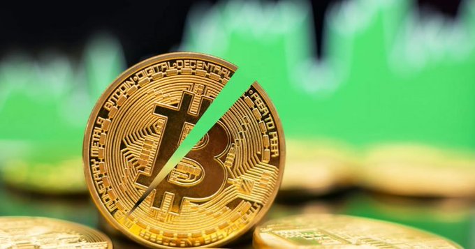 A gold Bitcoin token, visibly cracked in half, sits in the foreground against a backdrop of blurred green stock-chart patterns, visually symbolizing the upcoming Bitcoin halving event discussed in the post, which highlights the approaching 90,000 blocks until the next supply cut.