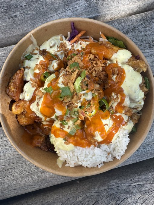 A paper bowl filled with white rice topped with fried chicken pieces, drizzled with orange and white sauces, garnished with green onions and crispy shallots, served on a wooden table.