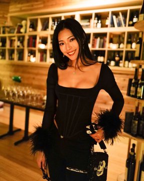 A woman in an elegant black corset-style dress with feathered cuffs poses in a wine cellar-style room lined with bottles, fitting the post's shoutout to the "Ladies of BAYC" with a stylish merch offering.