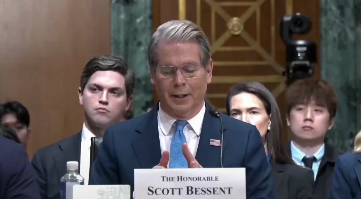The image shows a man giving a speech at a press conference, standing behind a podium while holding up a piece of paper that reads "Scott Bessent". The room has a green wall with a gold design, and there are other people present in the background who appear to be listening to the speech. The post also mentions that the US Treasury Secretary is looking forward to working with lawmakers on the Strategic Bitcoin Reserve.