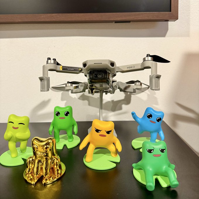 A DJI Mini 2 drone displayed on a stand surrounded by a collection of colorful cartoon figurines in green, yellow, blue, and gold finishes, all with expressive faces posed on green leaf bases.