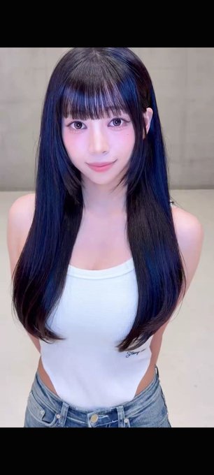 A young woman with long dark hair and bangs, wearing a white tank top and jeans, smiles softly at the camera.