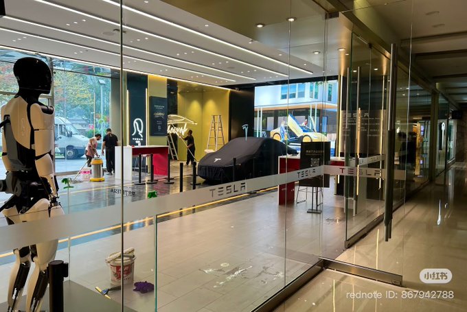 A photograph taken through a glass window shows a Tesla showroom with the word 'TESLA' across the glass; inside, there is a humanoid robot figure, covered vehicles, and a ladder.
