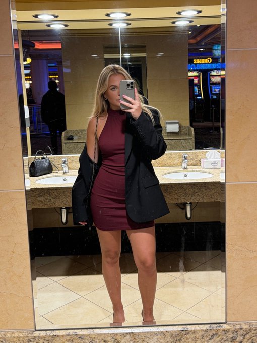 A blonde woman in a burgundy mini dress and black blazer takes a mirror selfie in what appears to be a casino restroom, with slot machines visible in the background. The image ties into the post by showcasing the casino setting that hints at her poker winnings, inviting guesses on her take.