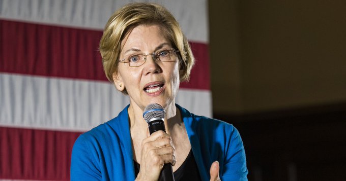 Elizabeth Warren, wearing a blue cardigan and glasses, speaks into a microphone with an American flag draped in the background, as she faces criticism from Cardano's founder for her stance on cryptocurrency regulation.