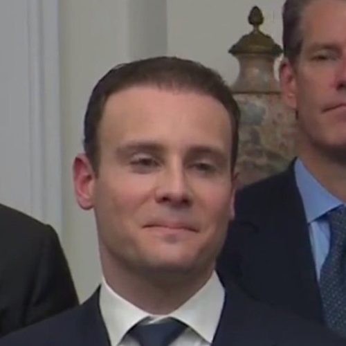 A close-up of a man in a dark suit with a subtle smile, with another man partially visible in the background.