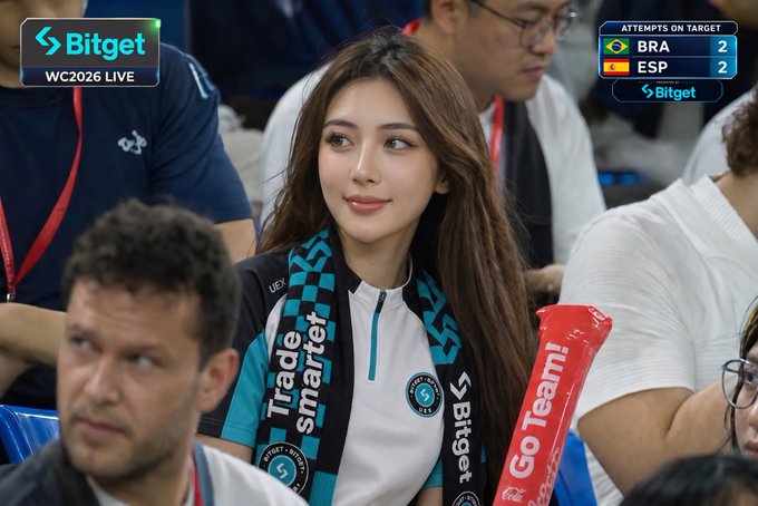 The image features a group of people sitting together during a football match. One person appears to be a woman wearing a scarf with black and blue colors, likely cheering for her team. The scene shows various other spectators seated around them. A laptop can also be seen in the background, possibly displaying information or statistics about the match.