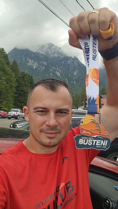 The image features a man holding up his race medal, which has the word "Ultra" written on it. The man appears to be posing for a photo with the mountain range visible behind him. The caption of the image reads, "On Saturday I ran at Busteni Ultra Race, 29 km. It was Ultra not just Trail It was difficult, but I filled my soul with peace. I burned a lot of energy #STEPN but the only problem on the mountain was that there was no internet signal. What's next? Carpathia Trails on July".