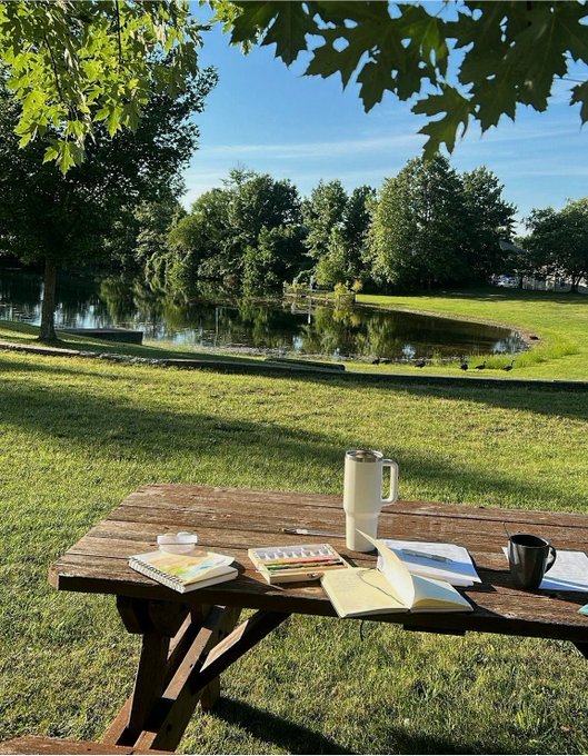 A wooden picnic table in a grassy park by a pond, set up with a pitcher, notebook, watercolor palette, and coffee mug for outdoor painting.