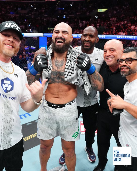 A bearded fighter with tattoos, wearing a keffiyeh and UFC gloves, celebrates victoriously with his team inside the octagon at UFC 331, with "New Amsterdam Vodka" and "Crypto.com" logos visible. The post highlights the camaraderie of having his corner by his side during the win.
