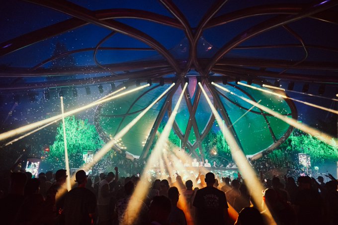 A concert crowd silhouetted against bright, radiating stage lights and green laser beams inside a large venue with a complex structural ceiling.