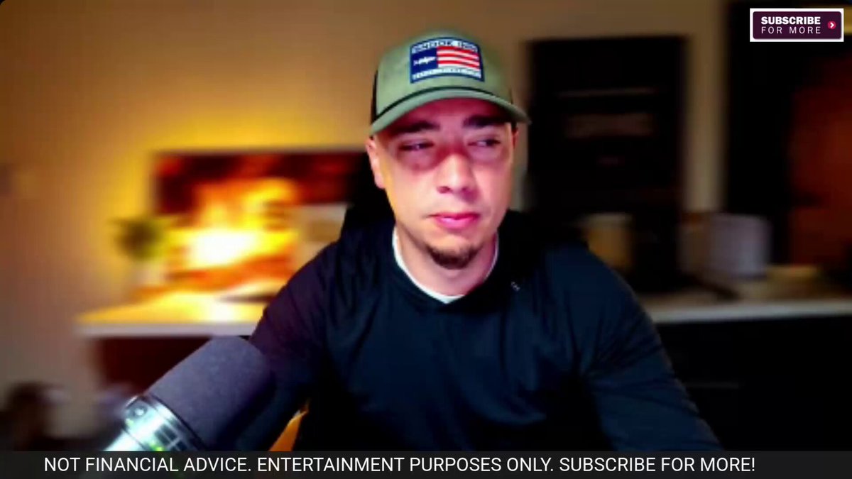 In the image, there is a man wearing a green hat sitting behind a microphone. He appears to be a TV host or presenter participating in an interview or discussion. The room has a wooden desk with various items scattered around it, including a potted plant and a cup on the surface.
