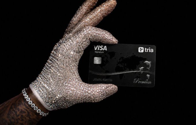 A hand wearing a sparkly, rhinestone-encrusted glove and a tennis bracelet holds a black Visa Signature credit card branded with the "tria" logo, displaying the name "JOHN SMITH" and the word "Premium."