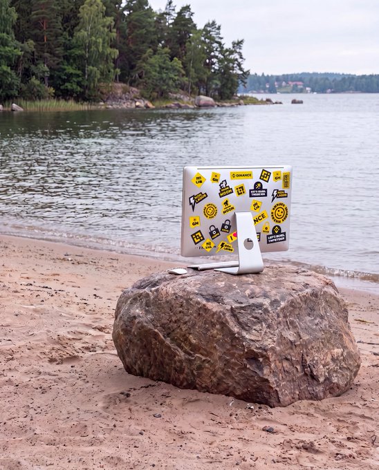 A silver laptop covered in developer and tech-themed stickers sits open on a large rock at the edge of a sandy beach, with calm water and forested shoreline in the background—capturing a serene "Friday setup" for remote work in nature.