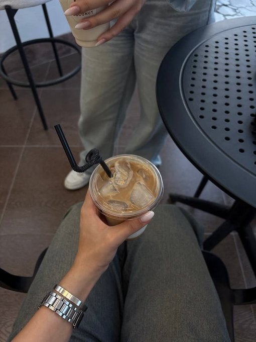 A first-person perspective of someone holding an iced coffee in a plastic cup while seated, with another person standing nearby holding a coconut drink.