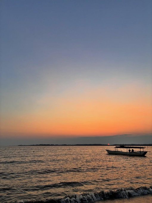 A serene sunset view over the ocean with a small boat carrying people silhouetted against the orange horizon.
