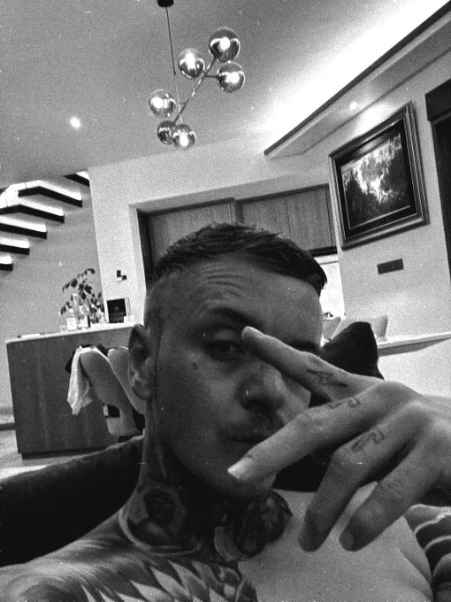 A black and white selfie of a heavily tattooed man making a hand gesture, with a modern interior featuring a spherical chandelier and a framed picture in the background.